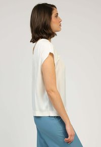 Blouse blanche à manches courtes avec une coupe décontractée et une texture douce. L'ourlet est légèrement incurvé, associée à un pantalon bleu.