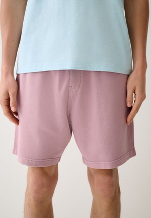 Persoon draagt een lichtblauw overhemd en roze korte broek, staand met handen ontspannen langs het lichaam.