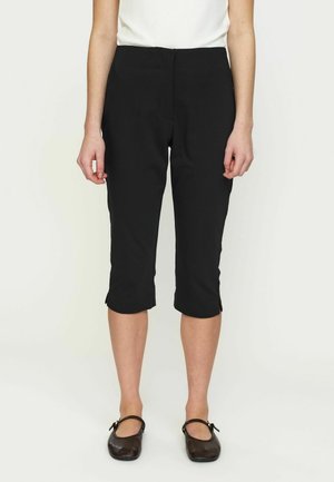 Kobieta w czarnych spodniach 3/4 i czarnych butach Mary Jane, stoi na tle jednolitego jasnego tła.