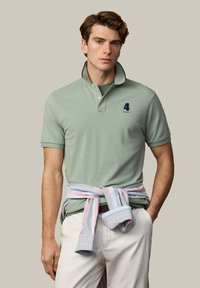 Mladý muž v svetlo zelenom polokošeli, pruhovanej pastelovej košeli zviazanej okolo pása a bielych nohaviciach, stojaci s jednou rukou v vrecku.