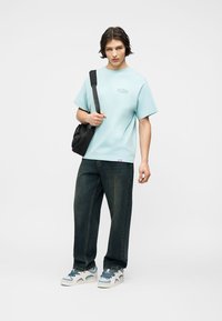 Νεαρός άνδρας που φοράει ανοιχτό μπλε φαρδύ T-shirt, σκούρο τζιν, αθλητικά παπούτσια και μαύρη τσάντα χιαστί, στέκεται μπροστά σε απλό λευκό φόντο.