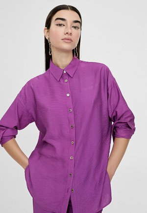 Camisa de botón morada con un acabado texturizado, que presenta un cuello clásico, mangas largas remangadas y bolsillos frontales.