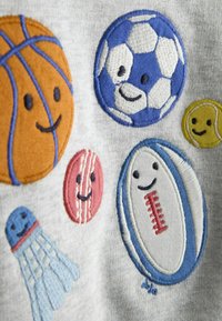 Sivá tkanina s vyšívanými športovými ikonami: basketbal, futbal, ragby a bedminton, každá s usmievajúcimi sa tvárami v žiarivých farbách ako oranžová, modrá, žltá, červená a ružová.