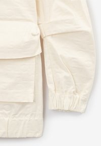 Krémově zbarvená bunda z texturované látky s jemným vzorem, elastickými manžetami a přední kapsou s klopou.