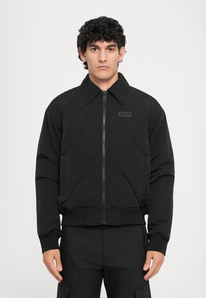 Black quilted jacket with a zipper, featuring a collar and ribbed cuffs. Includes a logo patch on the chest and a cropped design.
