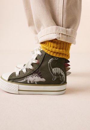 Barn iført olivengrønne høje sneakers med dinosaurprint og stofspidser på hælen, kombineret med gule strikkede sokker og beige bukser.