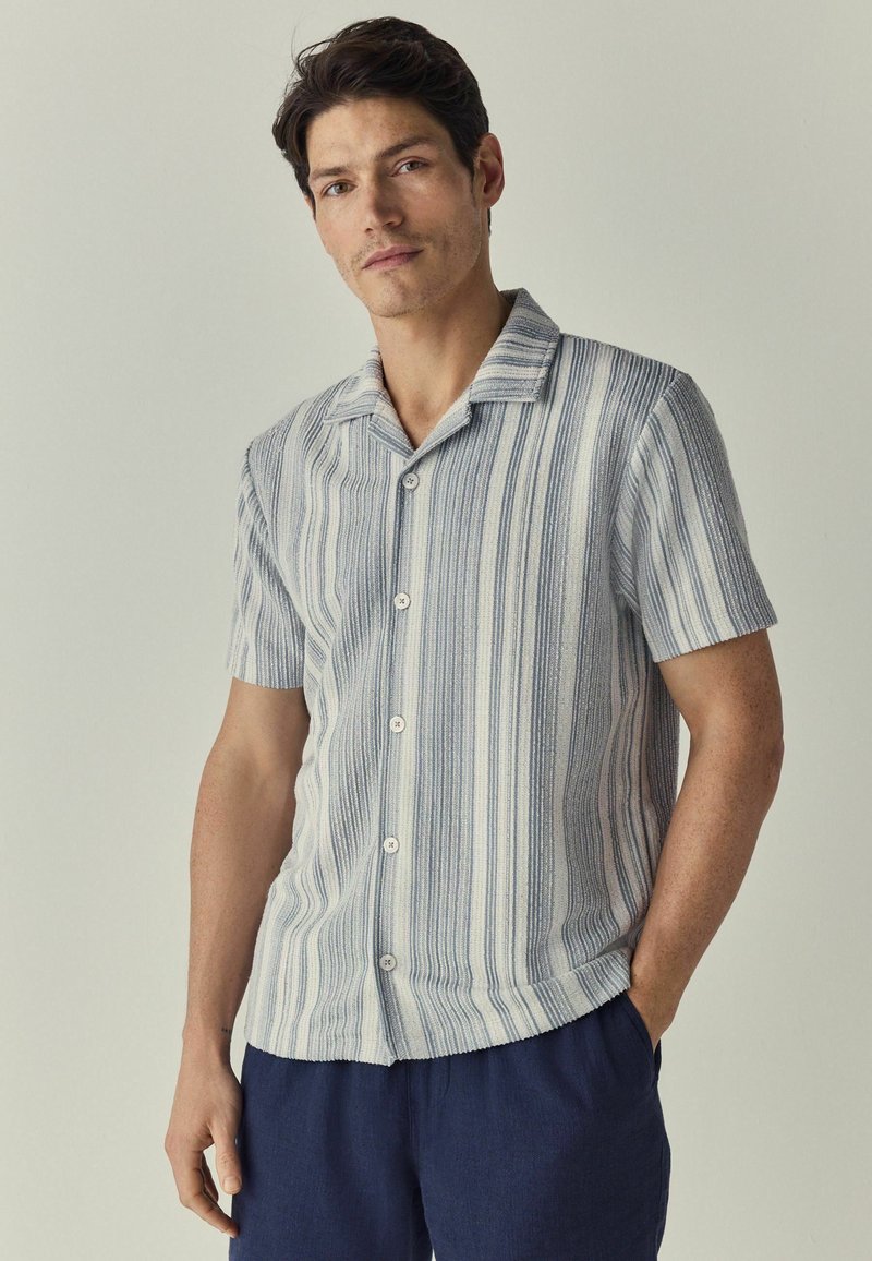 Chemise à manches courtes à rayures blanches et bleues, fabriquée en tissu texturé, avec un col et cinq boutons sur le devant.