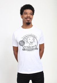 Bílý bavlněný tričko s grafickou ilustrací usmívající se krávy. Text zahrnuje "La Vache qui rit" a dekorativní prvky v šedé barvě.