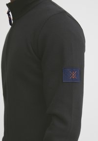 Mustat vetoketjulliset villatakit, joissa on korkeampi kaulus. Hihoissa sininen patch oranssilla embleemi- ja kuvioinnilla. Karhea kangas, jossa on ribattu yksityiskohta.