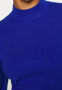 Maglione a collo alto lavorato a maglia blu con una texture liscia e un design aderente. Presenta un colletto alto e dettagli a coste.