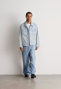 Man wearing light blue textured jacket, white shirt, loose blue jeans, and black shoes walking on gray floor against white wall.