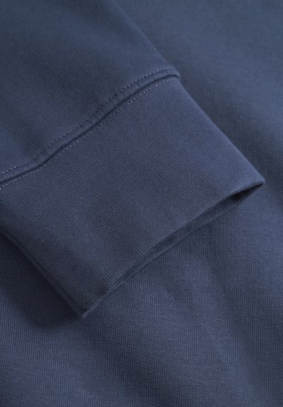 Tissu de sweatshirt bleu marine avec un poignet côtelé, présentant une texture lisse et des coutures soignées. Le poignet est replié, révélant des lignes épurées.