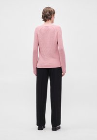 Ανοιχτό ροζ πουλόβερ πλεκτό με υφή και ραγκλάν μανίκια, φορεμένο με μαύρα παντελόνια ίσιας γραμμής και σκούρα υποδήματα.