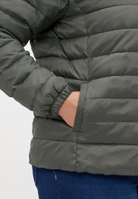 Olivengrønn pufferjakke med horisontale quiltede seksjoner, elastiske mansjetter og sidelommer. Materialet har en jevn, blank finish.