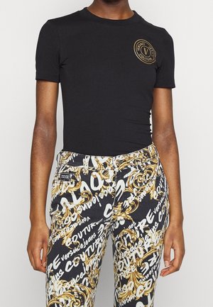Person iført en sort Versace Jeans Couture T-shirt og mønstrede bukser med gyldne barokdesigns og hvid skrift.