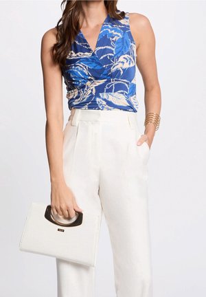 Donna che indossa un top smanicato blu con motivo e pantaloni bianchi a vita alta, che tiene in una mano una borsa bianca con dettagli dorati.