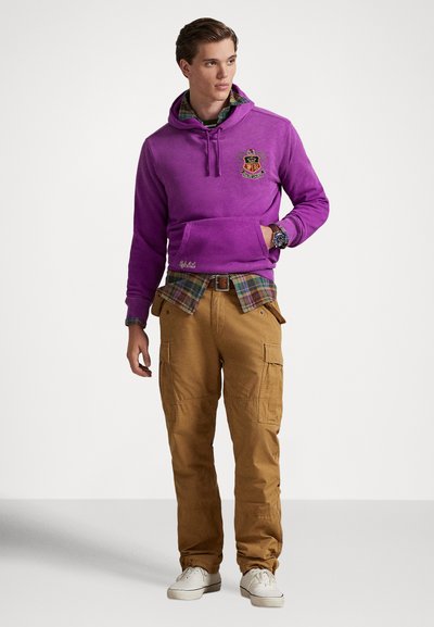 Fialová mikina s kapucňou s logom, vrstvená cez károvanú košeľu. Béžové cargo nohavice s viacerými vreckami a biele tenisky. Hladká textúra, pohodlný strih.
