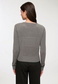 Maglione grigio lavorato a maglia con orlo e polsini a costine, adornato con piccoli elementi decorativi sulle maniche, visto da dietro. Pantaloni neri a righe.