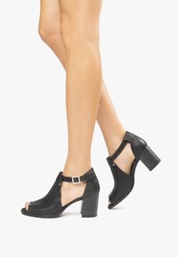 Black peep-toe heeled shoes with a textured upper, ankle straps, and a chunky heel. Buckle detail and open sides enhance design.