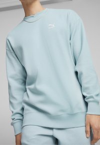 Person som bär en ljusblå texturerad Puma-tröja och matchande byxor, visar torso och en del av halsen och handen.