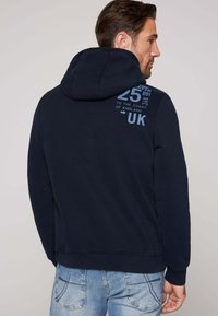 Sudadera con capucha en color navy que presenta un bolsillo canguro en la parte frontal, puños acanalados y detalles de impresión en la parte trasera. Tejido suave con un ajuste relajado.