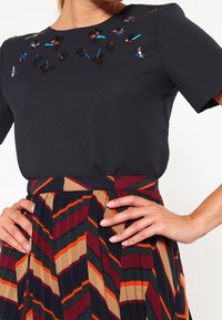 Top nero con ricamo floreale sul collo, abbinato a una gonna plissettata con strisce diagonali multicolori in bordeaux, verde e arancione.