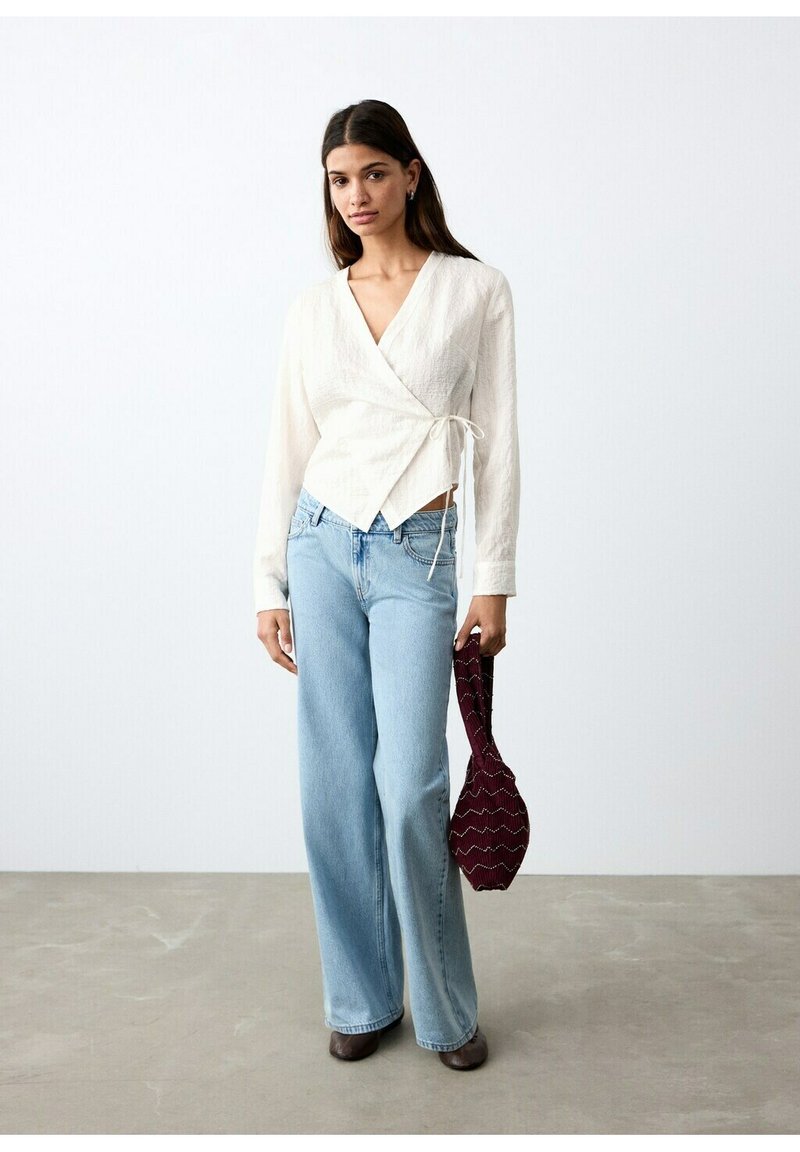 Femme portant un chemisier cache-cœur blanc, un jean pattes d'éléphant bleu clair, des chaussures marron, tenant un sac à main rouge foncé à motifs à ses côtés.