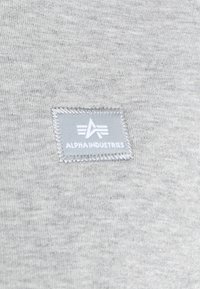 Tessuto grigio con una sottile texture. Presenta un'etichetta rettangolare cucita in un grigio chiaro, con la scritta "ALPHA INDUSTRIES" in bianco e il logo.