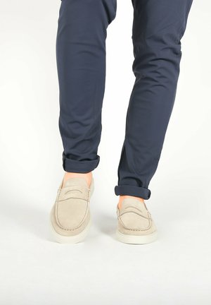 Beige suède loafers met een gestikt ontwerp, gecombineerd met donkerblauwe opgerolde broeken, die een gladde textuur en een lichte zool laten zien.