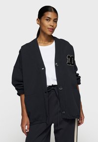 Schwarzer oversized Cardigan mit Knopfverschluss und Taschen, ausgestattet mit einer gestickten Zahl auf der Brust. Getragen über einem weißen T-Shirt.