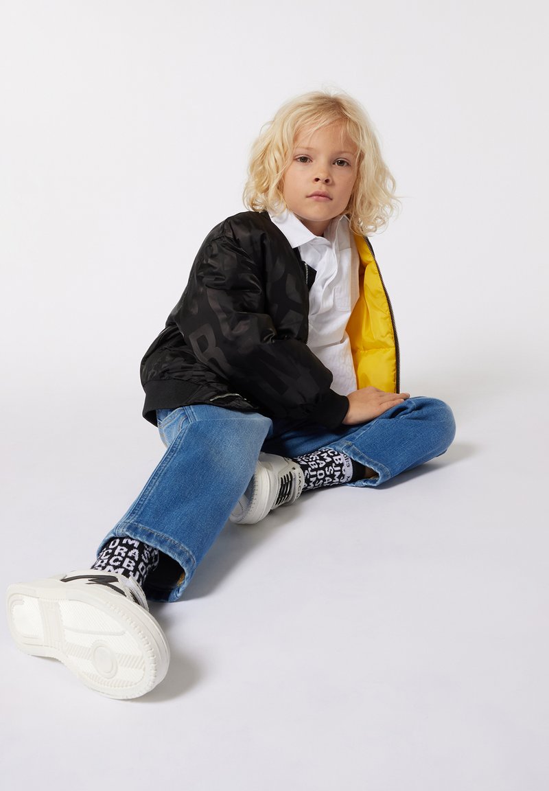 Zwarte bomberjack met gele voering, witte shirt, versleten blauwe jeans en zwart-wit gestreepte sokken gedragen met witte sneakers.