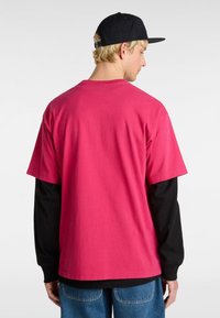 Kortærmet lyserød T-shirt lagdelt over en sort langærmet skjorte, kombineret med blå jeans. Stoffet ser blødt og afslappet ud.