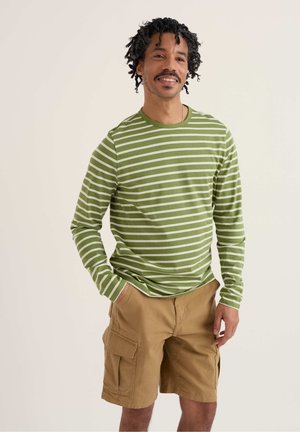 Uomo con capelli ricci che indossa una maglia a maniche lunghe a righe verdi e bianche e pantaloncini cargo beige, in piedi con le mani nelle tasche, sorridente.