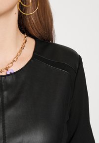 Svart lädertop med rund halsringning, subtila sömnadsdetaljer och ett guldhalsband med en lila accent.