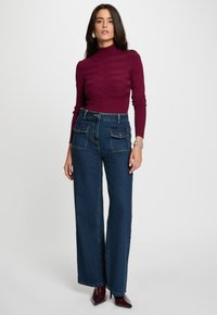 Burgundska turtleneck pulover s teksturnim uzorcima, uparen s tamnoplavo visokim hlačama širokih nogavica koje imaju prednje džepove i srebrne detalje.