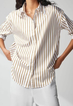 Femme portant une chemise boutonnée à rayures verticales blanches et marron clair avec les manches retroussées et un pantalon blanc, les mains posées sur les hanches.