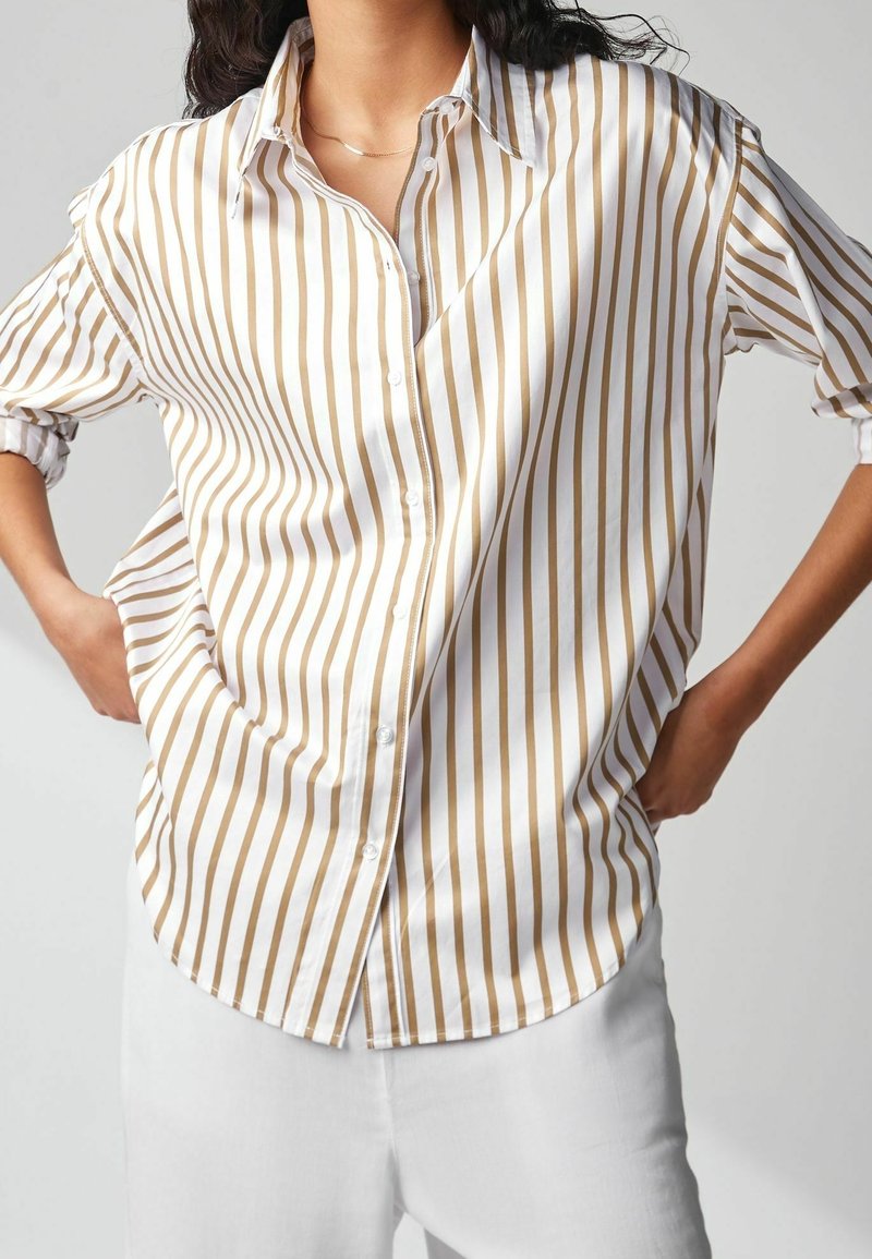 Femme portant une chemise boutonnée à rayures verticales blanches et marron clair avec les manches retroussées et un pantalon blanc, les mains posées sur les hanches.