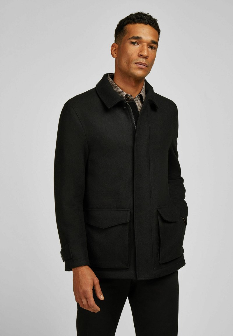 Man in een zwarte wollen jas met klepzakken en een geruit overhemd eronder, staande tegen een effen lichte achtergrond.