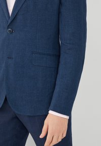 Mörkblå linnéblazer med en enkel knappstängning, slagkragar och en bröstficka. Manschetterna avslöjar en ljusrosa skjorta under.