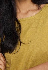 Haut à manches longues de couleur jaune moutarde avec une texture côtelée et un col large. Cheveux noirs partiellement visibles, bras croisés.