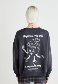 Čierna mikina s oversized siluetou. Obsahuje biely kreslený grafický motív cupcaku s rukami a textom "Šťastie je len cupcake ďaleko."