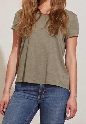 Kobieta ubrana w oliwkowozielony t-shirt z krótkim rękawem i niebieskie dżinsy, stojąca na jasnym tle z luźną postawą.