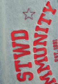 Ljusblå denimtyg med djärv röd broderad text och en stjärndesign. Broderiet är strukturerat med en upphöjd yta.