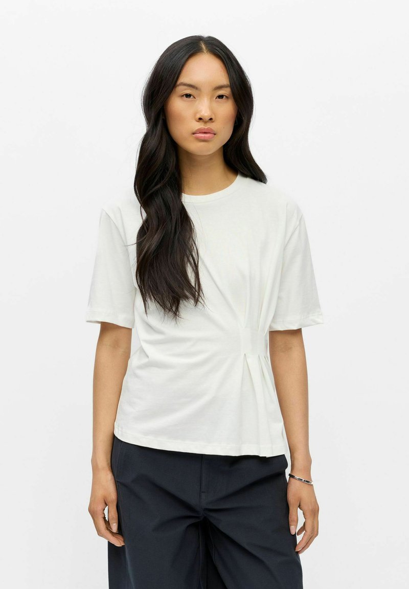 Mujer con cabello largo y oscuro, que lleva una camisa blanca de manga corta con detalle plisado y pantalones negros, de pie frente a un fondo blanco liso.
