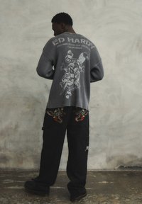 Maglione grigio a coste con grafica posteriore che presenta un teschio e una spada; pantaloni neri con ricami colorati di drago sulle tasche. Scarpe nere.