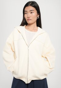 Krémová zipová mikina vyrobená z mäkkej tkaniny, s voľným strihom, oversized kapucňou a kontrastnými detailami šitia.