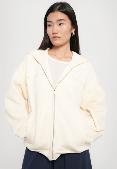 Cream-colored zip-up hoodie made of soft fabric, featuring a relaxed fit, oversized hood, and contrasting stitching details.