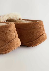 Barna velúr papucsok, puha, bolyhos belsővel, lekerekített orral és textúrált talppal a tapadás érdekében. A látható varrás részletgazdag megjelenést kölcsönöz.