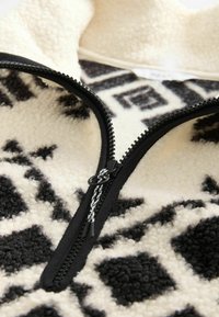 Gros plan sur un vêtement en polaire à motif noir et blanc avec une fermeture éclair noire partiellement ouverte et une tirette.