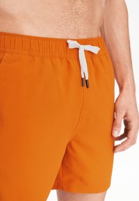 Oranžové plavky vyrobené z lehkého materiálu, s elastickým pasem a bílou tkaničkou s černými konci. Hladký povrch.